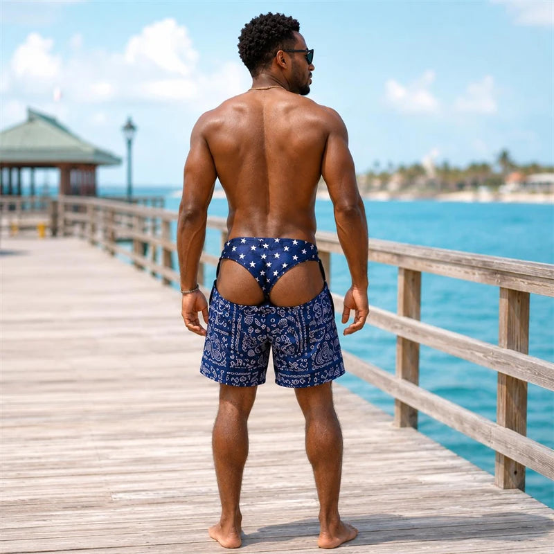 A man stands on a wooden pier with his back to the camera, wearing blue swim trunks with white star patterns on the back and a blue paisley pattern on the front, with a tropical ocean and resort in the background.