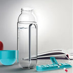 Clear glass water bottle with handle, blue cup, open book, and teal pill organizer on a light gray surface.