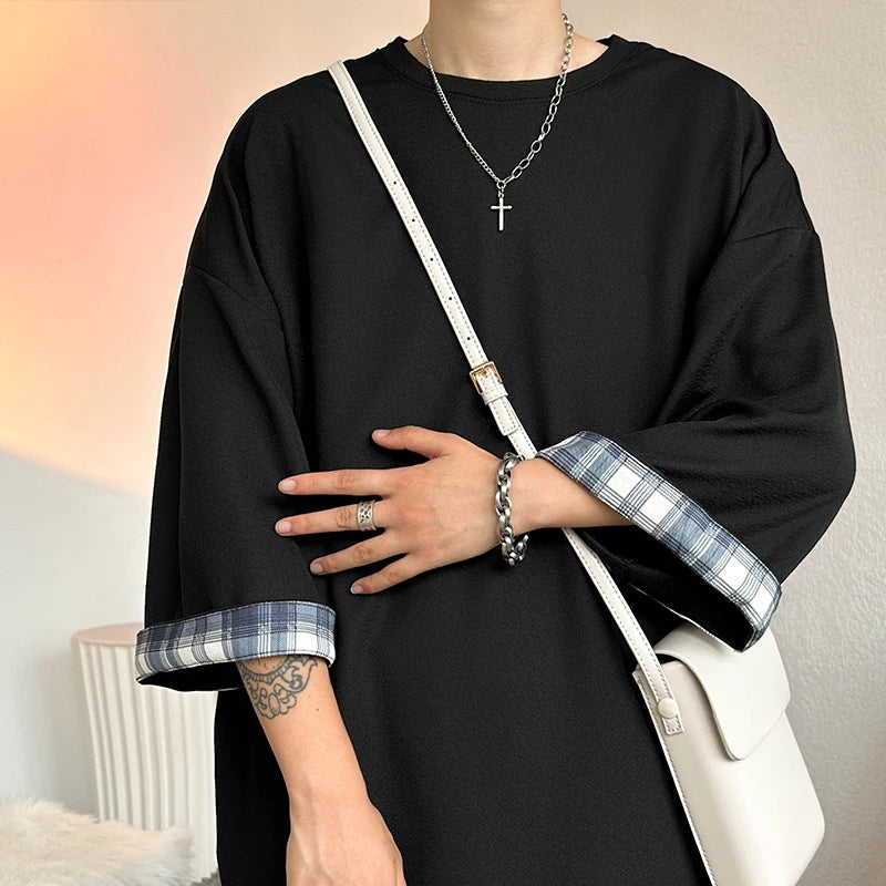 A person wearing an oversized black long-sleeve top with blue and white plaid turned-up cuffs, styled with a white crossbody bag and silver jewelry.