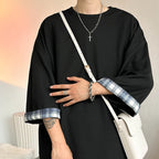 A person wearing an oversized black long-sleeve top with blue and white plaid turned-up cuffs, styled with a white crossbody bag and silver jewelry.
