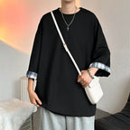 A person wearing an oversized black long-sleeve shirt with blue and white plaid rolled cuffs, paired with light gray sweatpants and a white crossbody bag, in an indoor setting.
