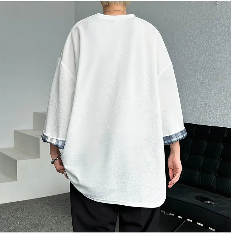 A person shown from the back wearing an oversized white long-sleeve top with contrasting plaid trim at the cuffs, paired with black pants in an indoor setting.