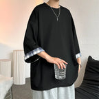 A person wearing an oversized black tunic-style top with rolled cuffs showing a blue-and-white plaid lining, paired with light gray sweatpants and a silver necklace, standing indoors and holding a clear glass.