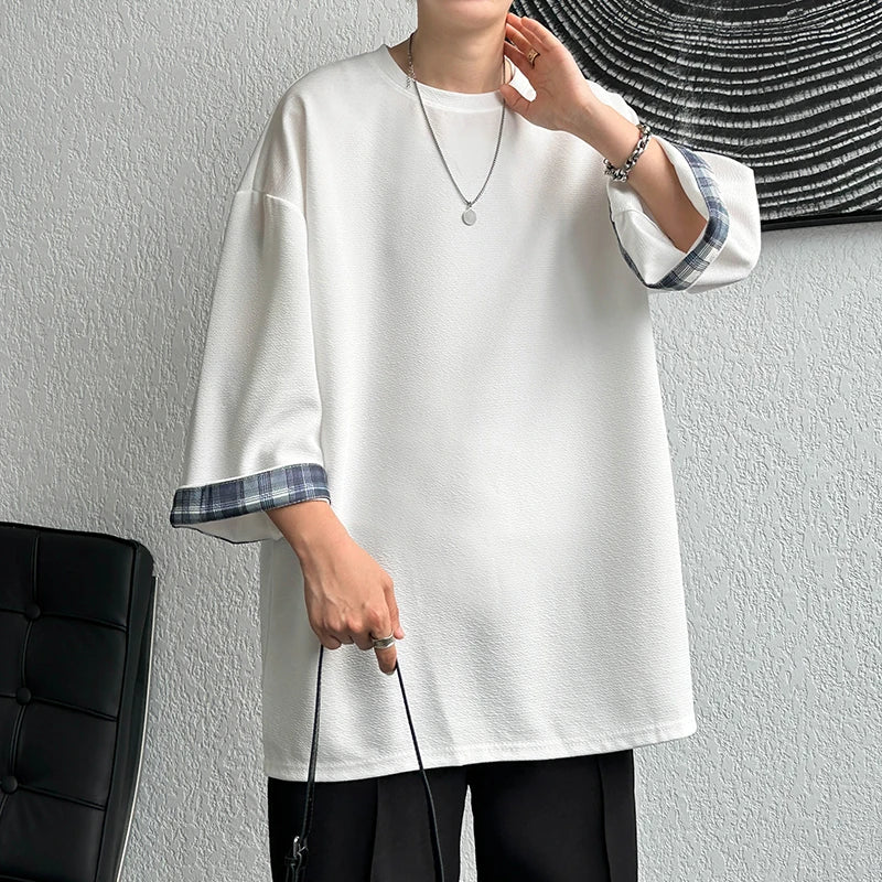 A person wearing an oversized white short-sleeve top with blue-gray plaid trim on the sleeve cuffs, paired with black pants and holding a black handbag against a textured light gray wall.