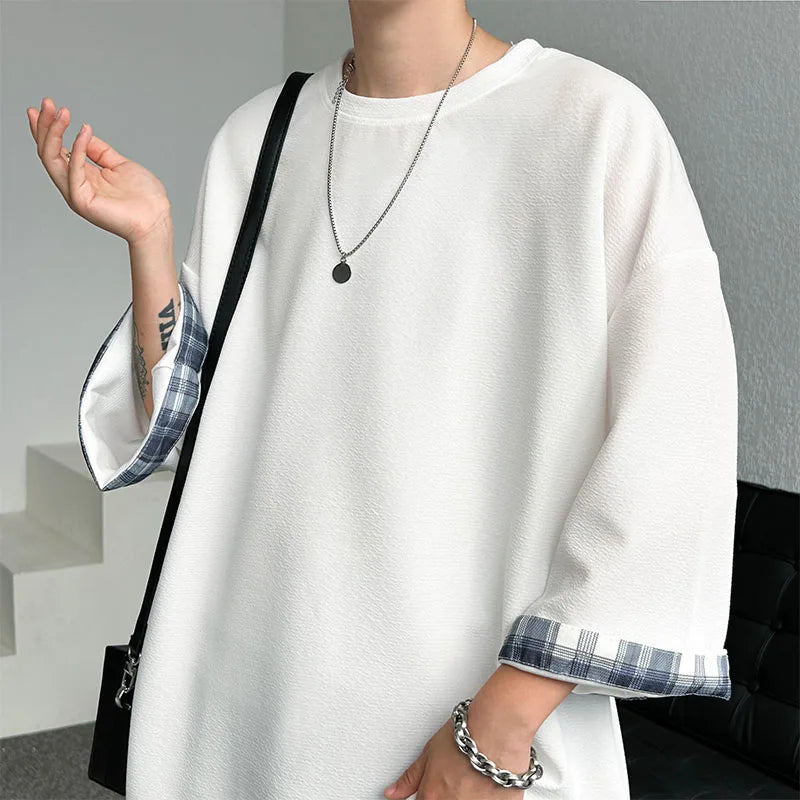 A person wearing an oversized white long-sleeve top with blue and white plaid turned-back cuffs, styled with layered silver necklaces and a silver chain bracelet, carrying a black shoulder bag.