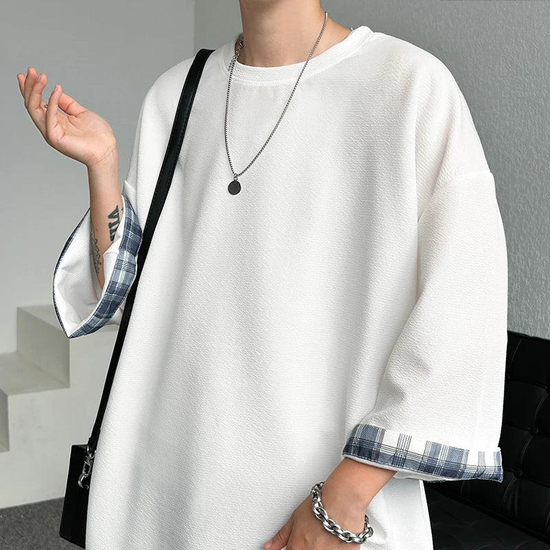 A person wearing an oversized white long-sleeve top with blue and white plaid turned-up cuffs, styled with layered silver necklaces and a silver chain bracelet, carrying a black shoulder bag.