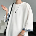 A person wearing an oversized white long-sleeve top with blue and white plaid turned-up cuffs, styled with layered silver necklaces and a silver chain bracelet, carrying a black shoulder bag.