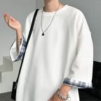 A person wearing an oversized white long-sleeve top with blue and white plaid turned-up cuffs, styled with layered silver necklaces and a silver chain bracelet, carrying a black shoulder bag.
