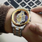 A close-up of a men's skeleton wristwatch with a gold and silver-tone metal case and link bracelet, worn on a person's wrist. The watch face is open, showing a detailed gold and silver mechanical movement.