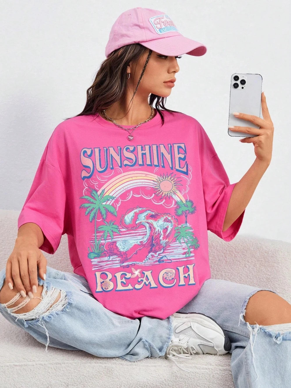 A woman sitting on a light gray couch takes a mirror selfie while wearing a bright pink graphic t-shirt with 'SUNSHINE BEACH' and a rainbow design, paired with ripped light blue jeans and a pink baseball cap.