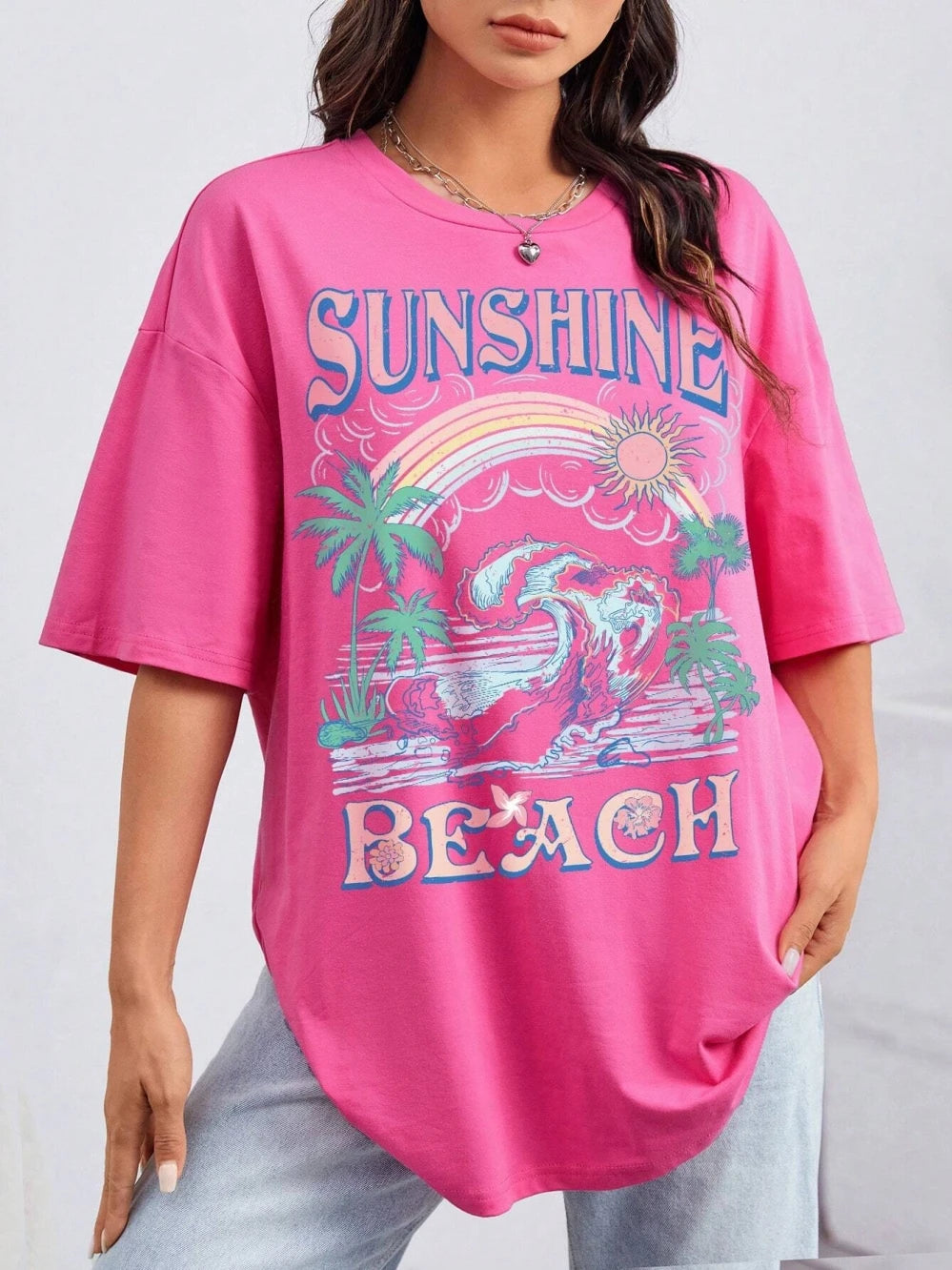 A woman wearing a bright pink oversized t-shirt with a colorful retro beach graphic that reads 'SUNSHINE BEACH' with palm trees and a rainbow.