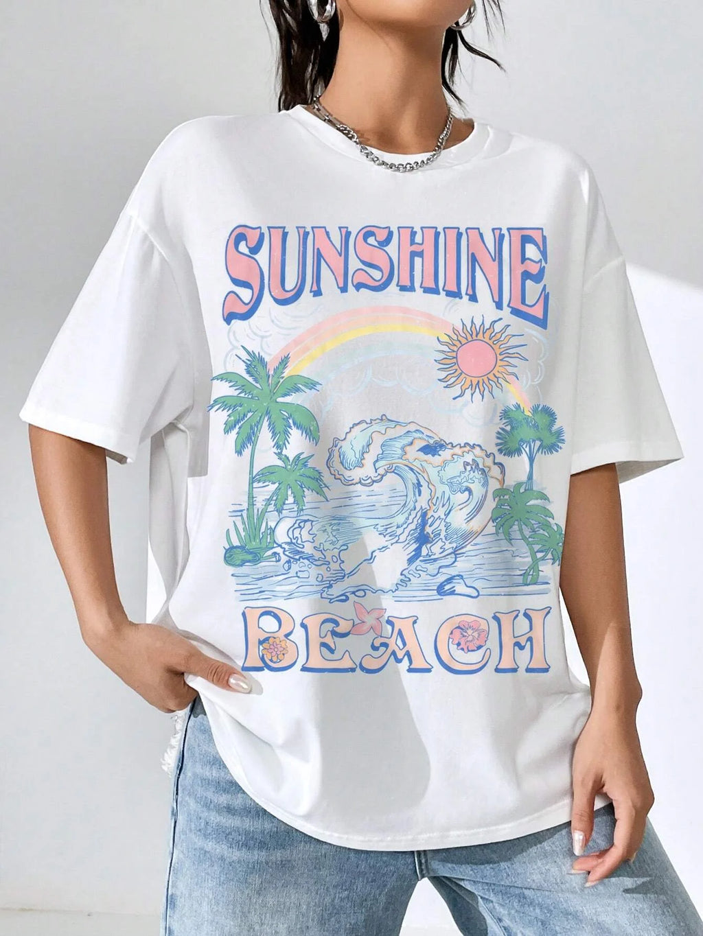 A woman wearing an oversized white graphic t-shirt with a colorful 'Sunshine Beach' design featuring palm trees, waves, and a rainbow, paired with light blue jeans.