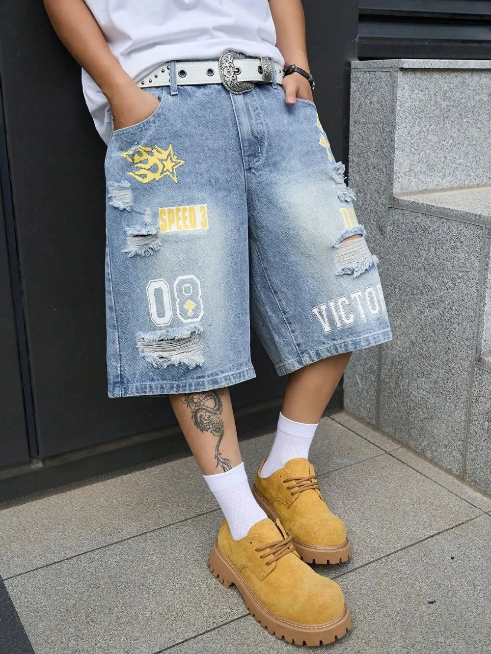 A person wearing light-wash distressed denim shorts with yellow and white graphic patches, a white t-shirt, white socks, and tan lace-up boots, standing on a gray tiled floor.