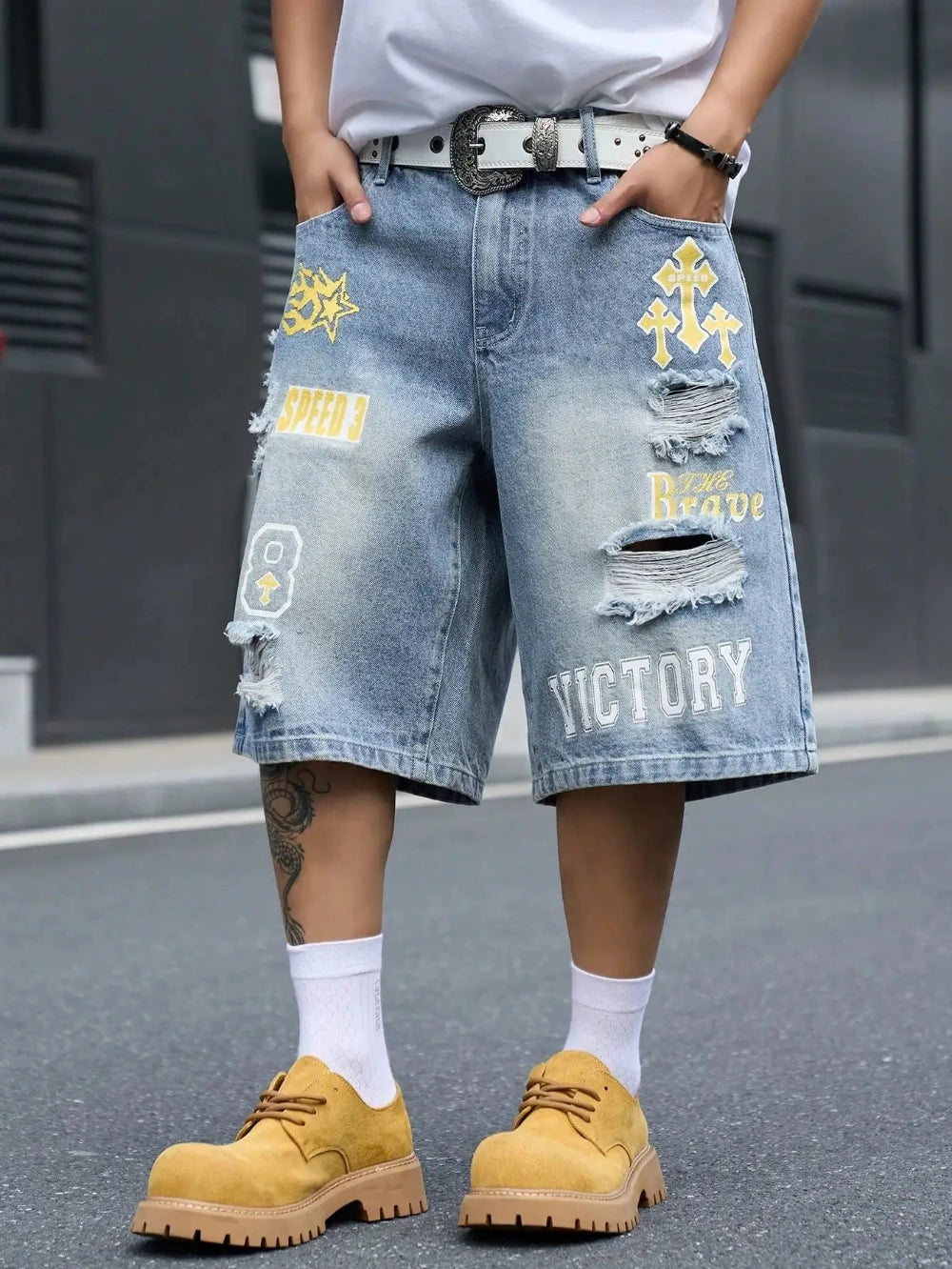 Light blue distressed denim shorts with yellow and white graphic patches, worn by a model with white socks and tan boots on a street background.