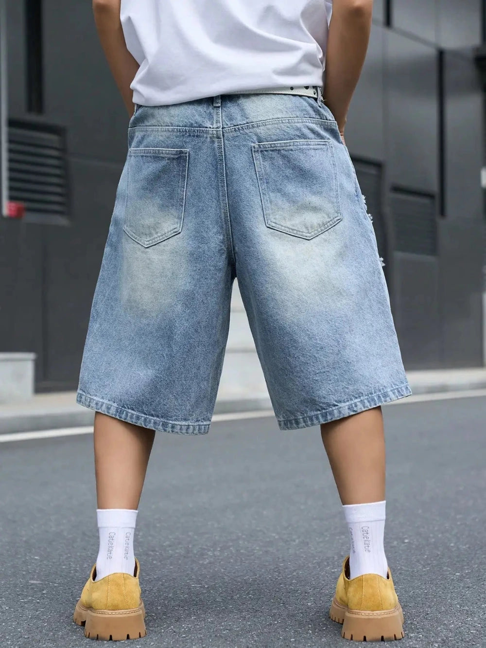 A person shown from the back wearing light-wash, wide-leg denim shorts with a faded wash, a white t-shirt, white socks, and mustard yellow slip-on shoes on an outdoor street.