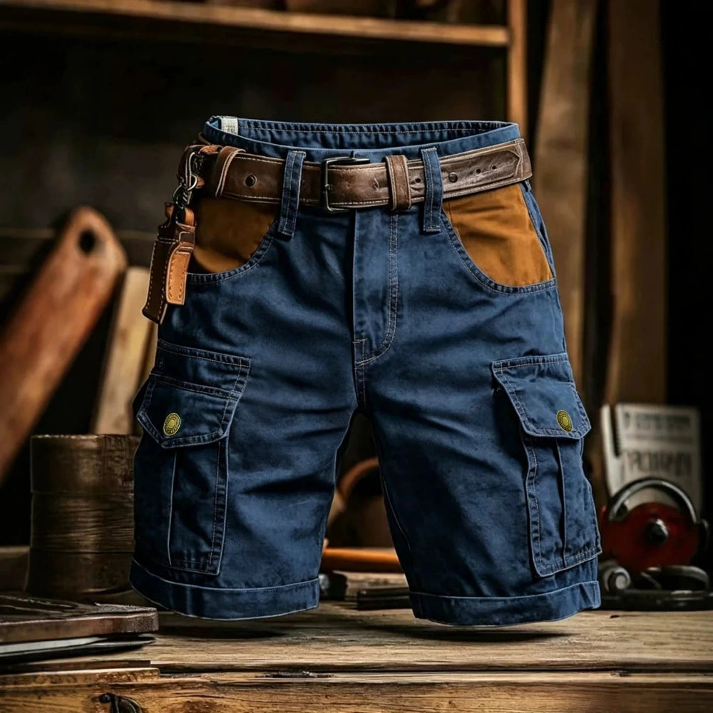 Blue denim cargo shorts with brown leather accents, including a matching belt, a leather key fob, and tan contrast panels on the pockets, displayed on a rustic wooden table.