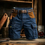 Blue denim cargo shorts with brown leather accents, including a matching belt, a leather key fob, and tan contrast panels on the pockets, displayed on a rustic wooden table.