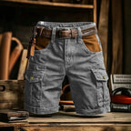 Gray cargo shorts with brown leather accents, including a matching belt, belt loops, and a brown leather patch on the pocket, displayed on a rustic wooden table.