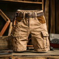 A pair of tan cargo shorts with a brown leather belt, displayed on a rustic wooden table with a workshop background.