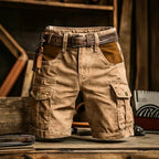 A pair of tan cargo shorts with a brown leather belt, displayed on a rustic wooden table with a workshop background.