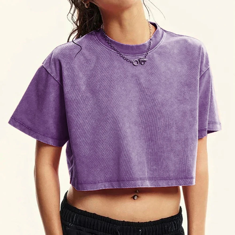 A person wearing a cropped short-sleeve ribbed T-shirt in a washed purple color, paired with black pants and a silver chain necklace, against a plain light background.