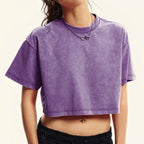 A person wearing a cropped short-sleeve ribbed T-shirt in a washed purple color, paired with black pants and a silver chain necklace, against a plain light background.