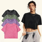 A model wears a black acid-wash cropped t-shirt, with four other color options (black, green, purple, pink) displayed on the left against a light cream background.