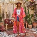 A woman stands barefoot on a patterned rug in a rustic outdoor patio setting, wearing a long pink and orange bohemian floral kimono robe over a white tank top and denim shorts.