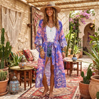 A woman stands barefoot in a bohemian courtyard wearing a long, open-front kimono with a purple and blue cloud print, a white tank top, denim shorts, and a wide-brim tan hat.