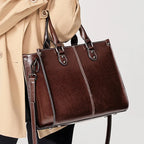 A dark brown leather tote bag with double top handles and a detachable shoulder strap, carried by a person wearing a beige trench coat against a light gray background.