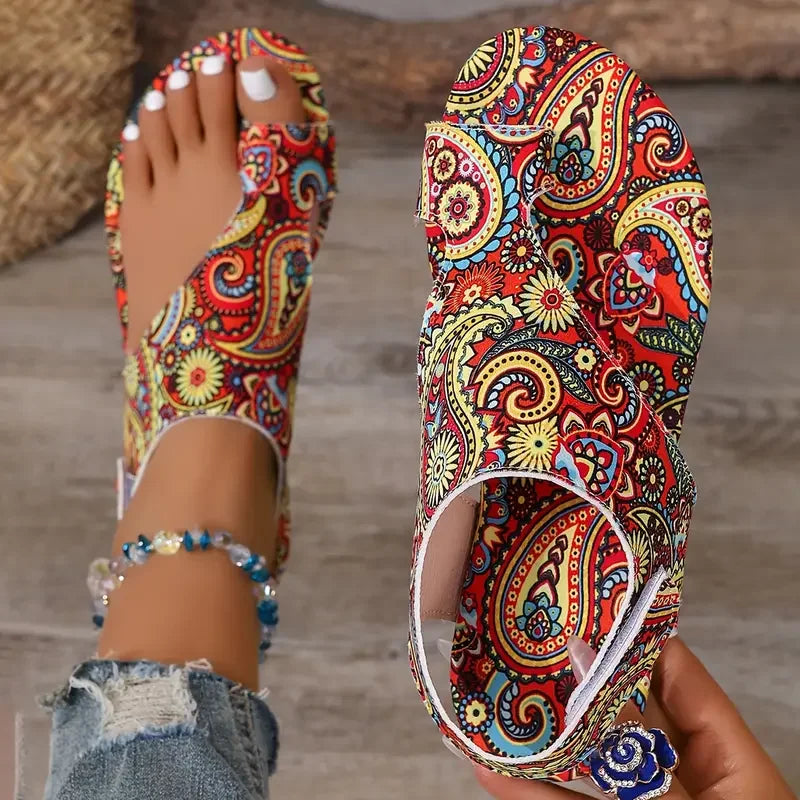 A pair of colorful open-toe flat sandals with a vibrant paisley pattern, shown worn on a foot and held in a hand against a neutral indoor background.