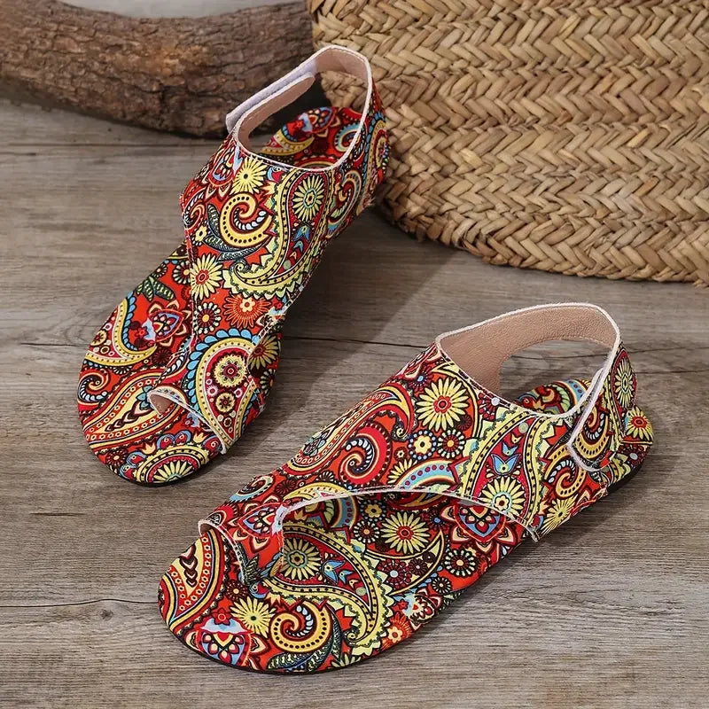 A pair of flat thong sandals with a vibrant, multicolored paisley pattern on a wood-textured floor, with a woven basket and log in the background.