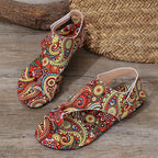 A pair of flat thong sandals with a vibrant, multicolored paisley pattern on a wood-textured floor, with a woven basket and log in the background.
