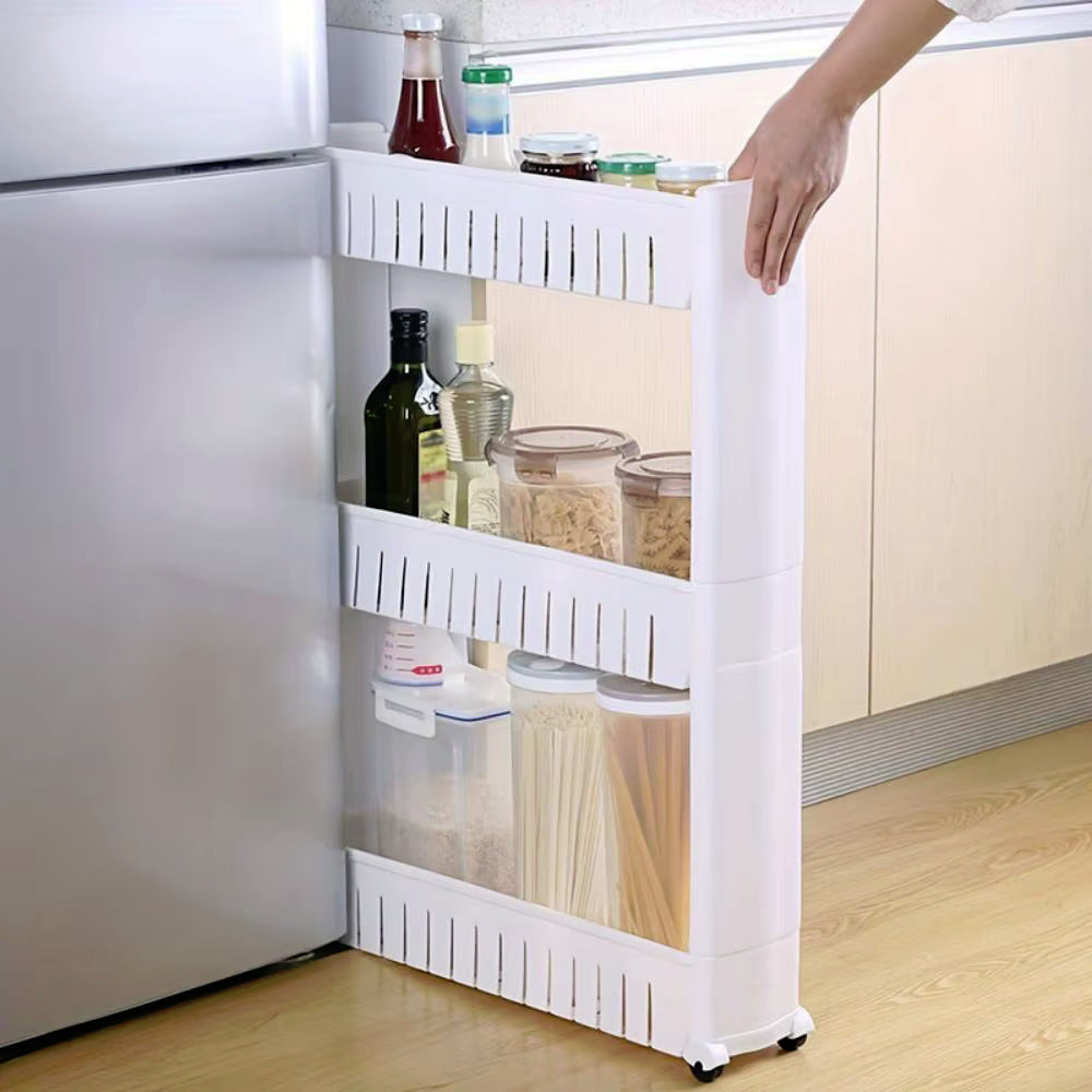 A white plastic under-counter refrigerator organizer shelf with three tiers, shown in a kitchen next to a stainless steel refrigerator. A person's hand is holding the handle on the right side to slide the organizer out.
