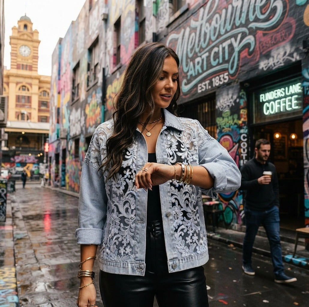 A woman wearing a light blue denim jacket with white floral embroidery stands on a wet city street in front of a graffiti-covered building. She is looking at her watch, with a coffee shop and another person visible in the background.